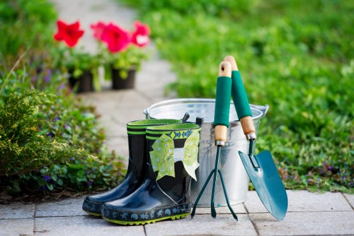 A close-up view of a garden pathway in an outdoor space, featuring a pair of black rubber wellington boots with a decorative green pattern, placed on a paved surface. Inside and around the boots are gardening tools including a small hand trowel with a blue metal blade and wooden handle, a pair of pruning shears with green handles, and a metal bucket, suggesting recent gardening activity. In the background, blurred vibrant pink and red flowering plants are visible, alongside lush green foliage and a well-maintained lawn area, indicating a healthy and cared-for garden environment. The scene is illuminated by natural daylight, highlighting the textures of the paved pathway, soil beds, and garden plantings, and reflecting a typical garden setup in Ilford for outdoor maintenance and landscaping services by Hedge Trimming Ilford, with a focus on lawn care and garden tidiness in a suburban setting.
