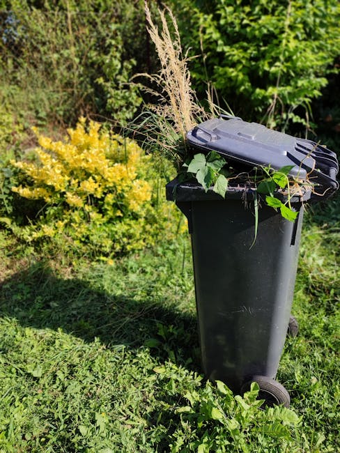 A black wheeled garden waste bin filled with trimmed plant debris, including long dried grass, leafy branches, and cut foliage, is positioned on a well-maintained garden lawn. Surrounding the bin are